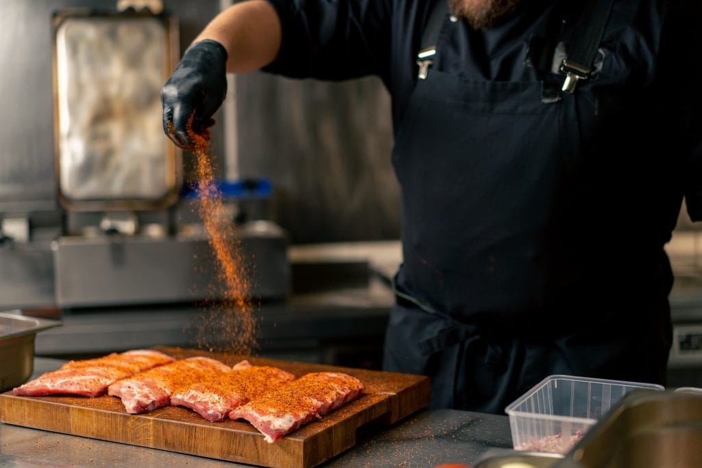 close-up in a professional kitchen in a chef in a black jacket peppers ribs that lie on wooden board