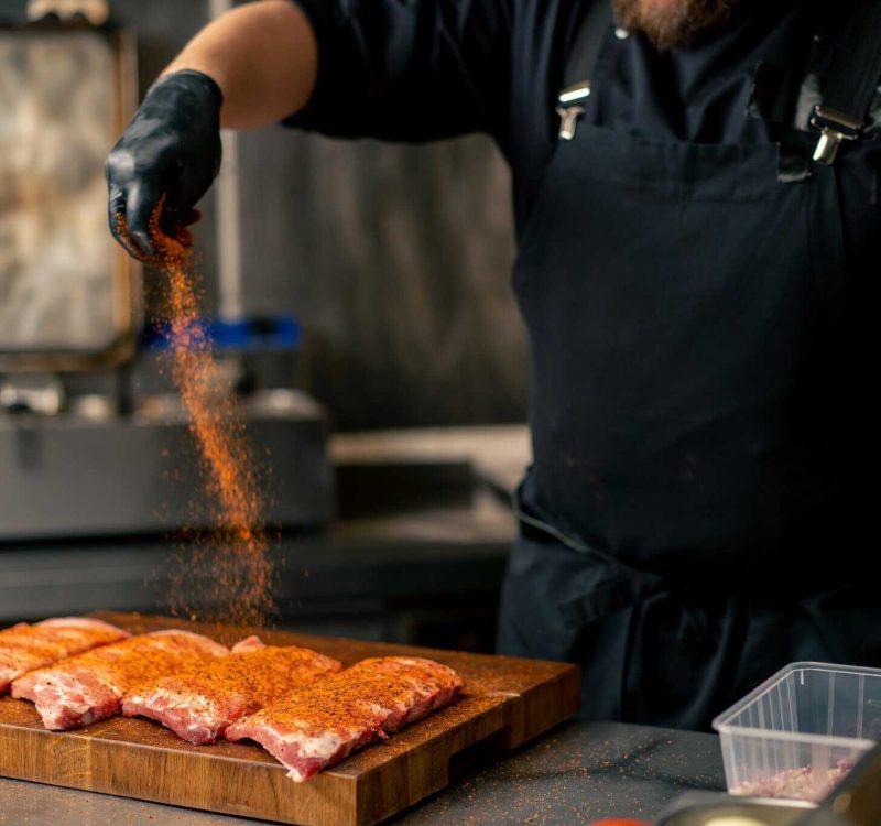 close-up in a professional kitchen in a chef in a black jacket peppers ribs that lie on wooden board